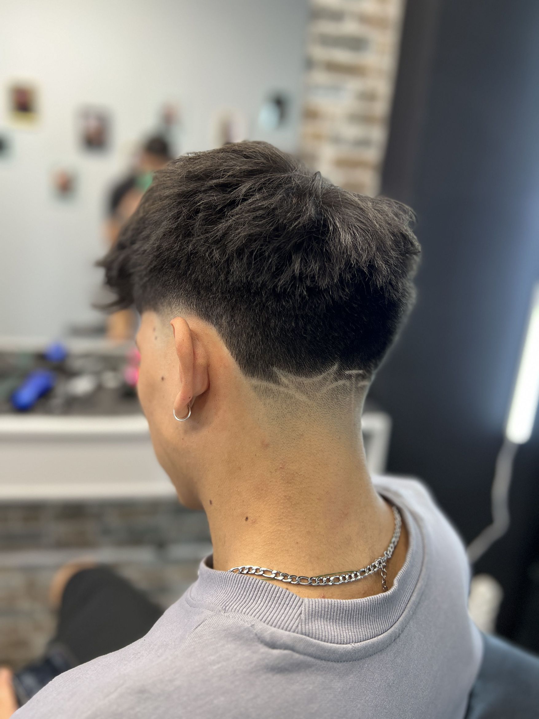 The back of a head with a textured top over a low taper and a shaved line behind the ear; a silver chain, a grey sweatshirt, and a brick wall and a barber's station out of focus behind.