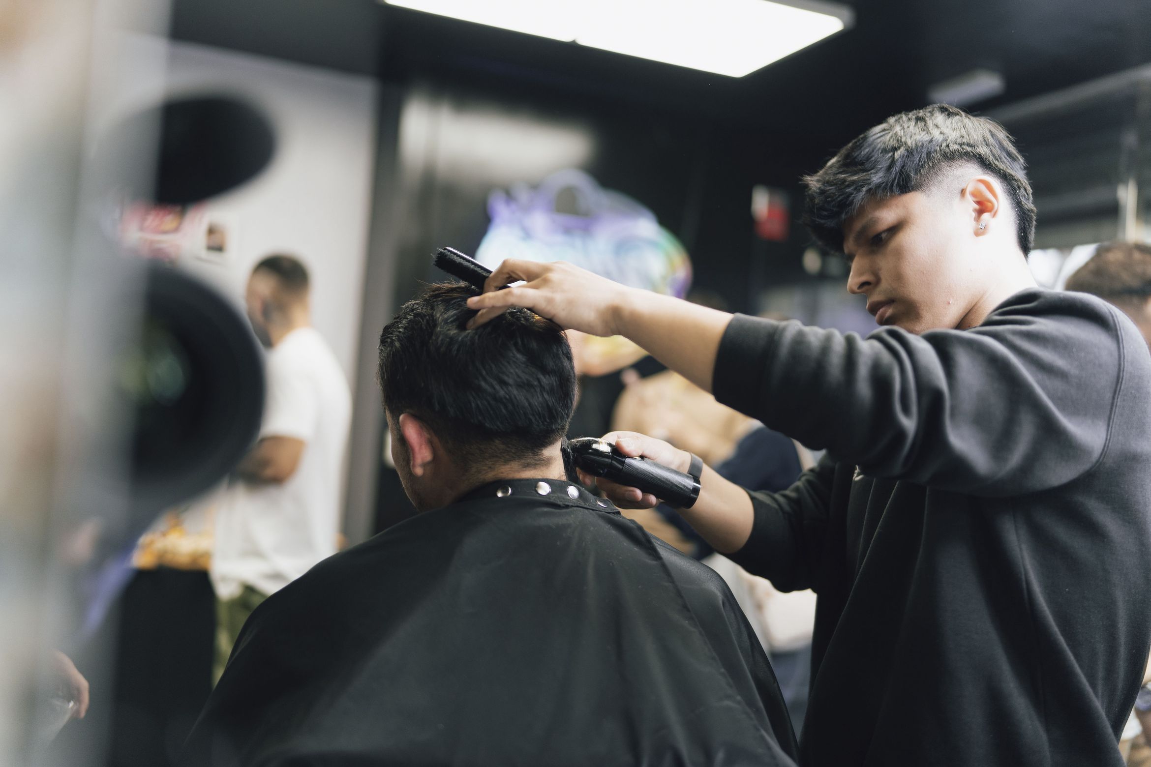 A barber in a black sweatshirt, seen in profile, running clippers over the nape of a client who is sitting with his back to the camera in a black cape; a mirror and another person out of focus behind.