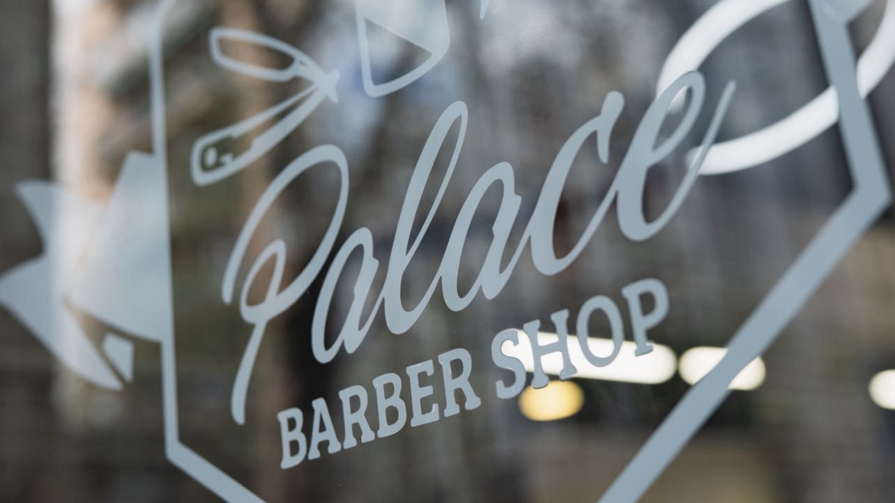 Close-up of the shop window: the crest etched on the glass with the Palace signature and the words BARBER SHOP, and the street out of focus beyond.