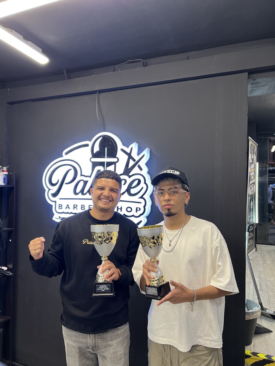 Dos personas con sudadera y camiseta sostienen sendos trofeos delante del rótulo retroiluminado Palace Barber Shop dentro del local.